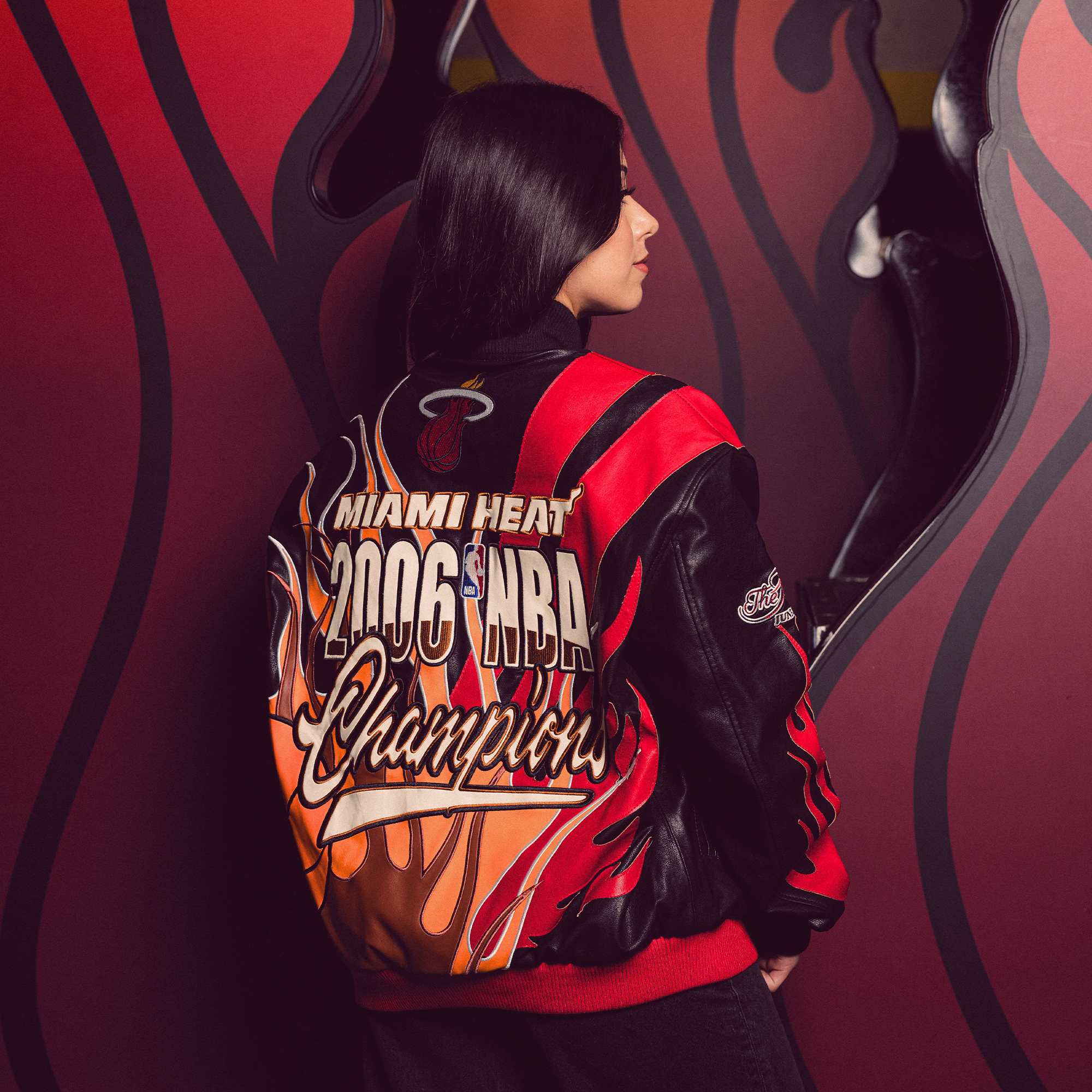 A woman faces away, wearing the Court Culture Miami HEAT x Jeff Hamilton 2006 Championship Legacy Jacket with basketball and flame graphics, standing beside a wall featuring a red and black flame design.