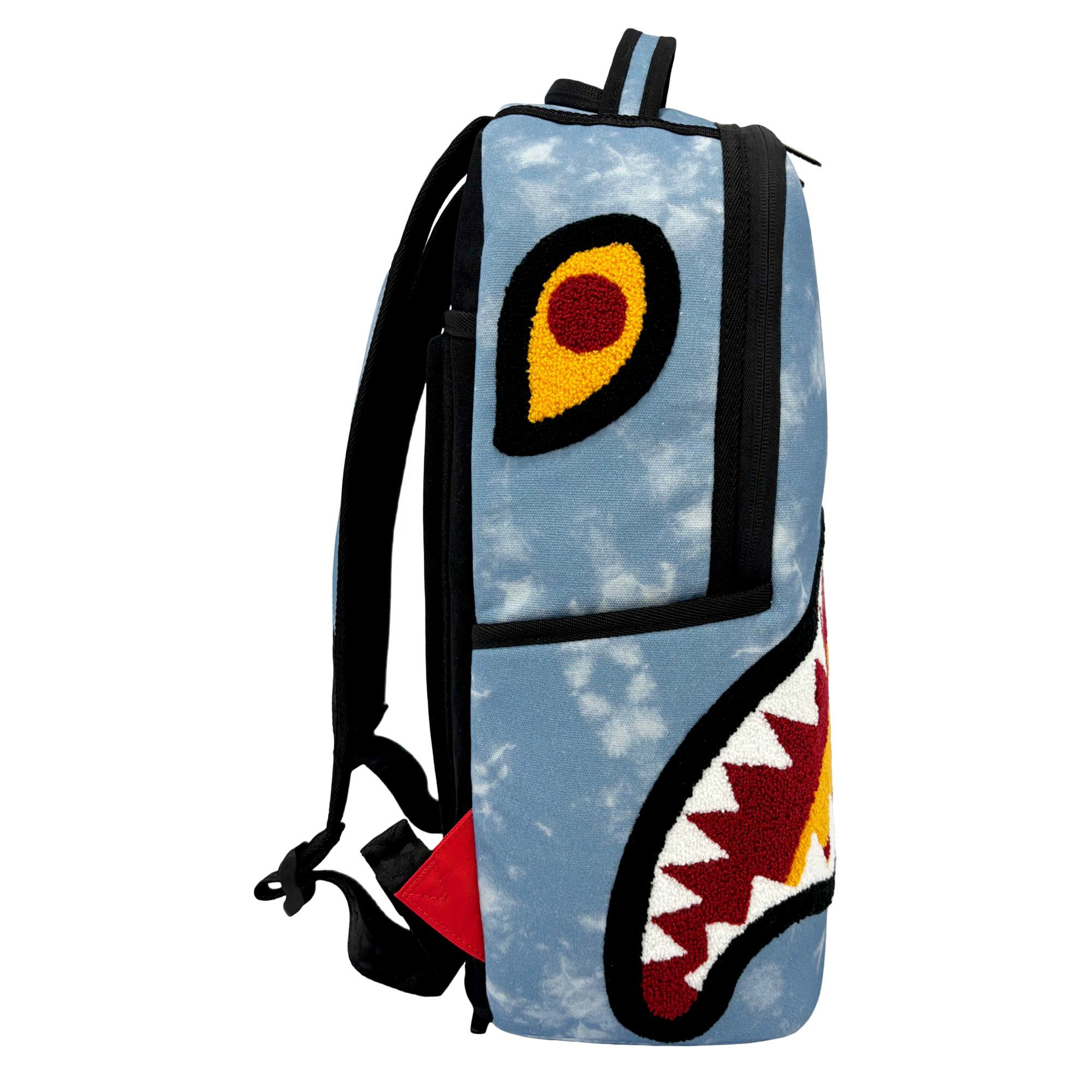 Introducing the Court Culture x Sprayground Miami HEAT Tie-Dye Jaw Backpack by Sprayground—a blue denim backpack with black straps, showcasing a stylized shark face design on the side, highlighted by a large open mouth with white teeth, a yellow eye accented with a red center, and an eye-catching tie-dye pattern background. This limited edition piece is finished with an embroidered logo for added detail.