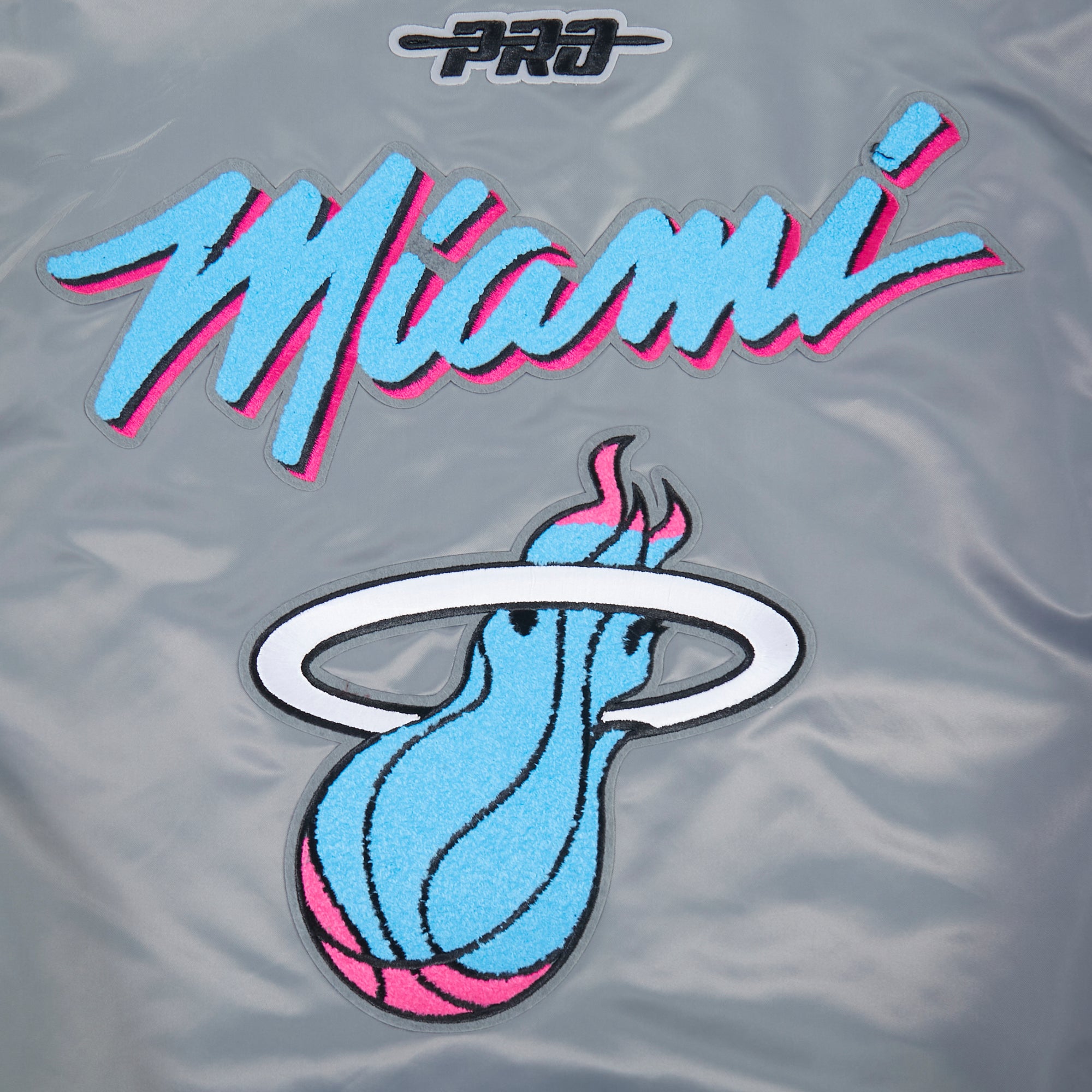 The Pro Standard Miami HEAT Original Vice Nights Satin Jacket features 'Miami' in bold blue and pink script with a blue flaming basketball logo below, outlined in white and black. This shiny gray polyester jacket is machine washable for easy care.