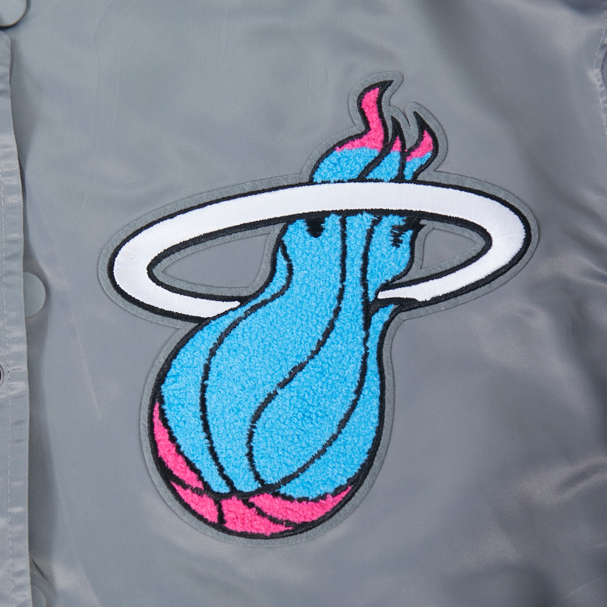 A close-up of the Pro Standard Miami HEAT Original Vice Nights Satin Jacket shows a colorful blue and pink flaming basketball patch with a white ring on gray polyester. This standard fit Pro Standard piece is stylish and machine washable.
