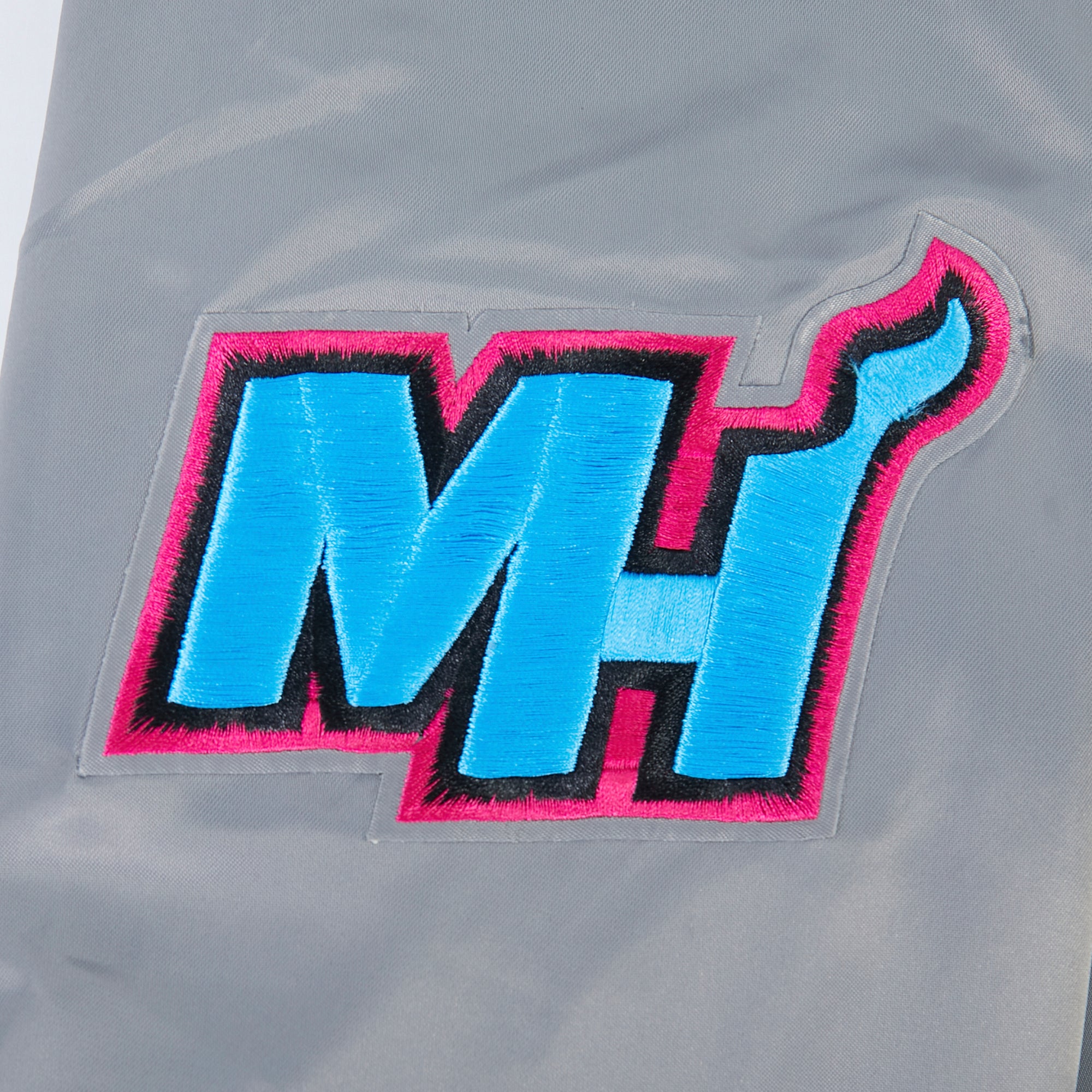 The Pro Standard Miami HEAT Original Vice Nights Satin Jacket features an embroidered patch with bold blue MH letters outlined in black and pink on gray polyester. The H curves into a hook shape. Machine washable for easy care.