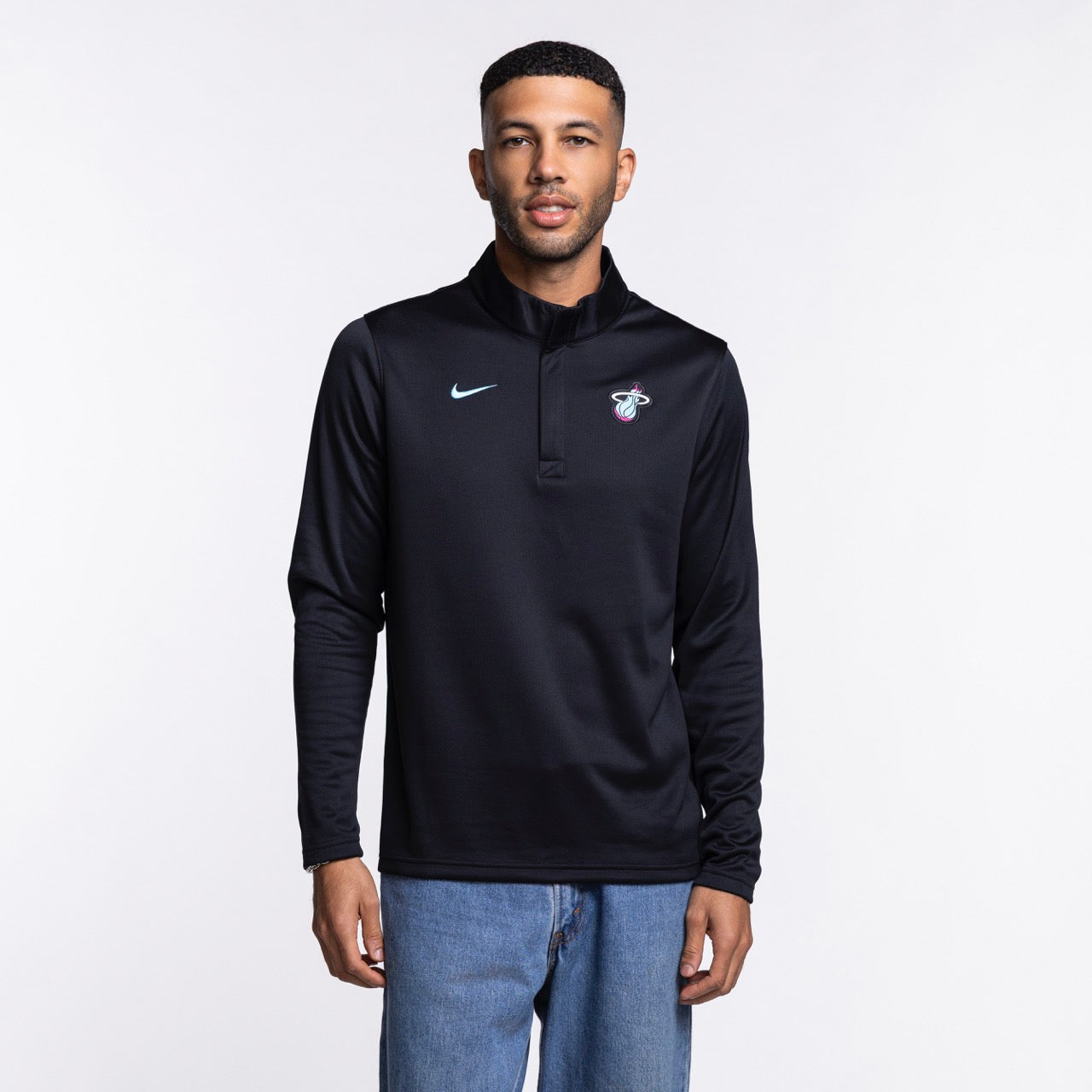 A man stands facing forward in a Nike Miami HEAT Original Vice Nights 1/4 Zip Jacket, featuring the team logo on the chest, paired with blue jeans. The black polyester pullover has a standard fit and is set against a plain white background.
