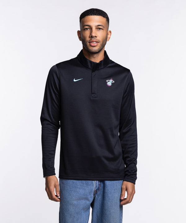 A man stands facing forward in a Nike Miami HEAT Original Vice Nights 1/4 Zip Jacket, featuring the team logo on the chest, paired with blue jeans. The black polyester pullover has a standard fit and is set against a plain white background.