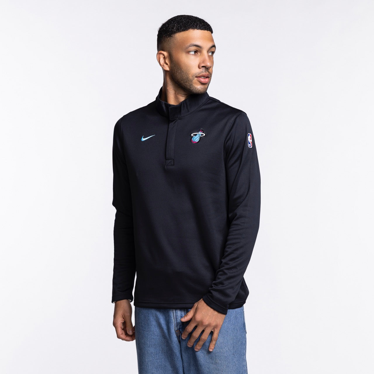 A man in a Nike Miami HEAT Original Vice Nights 1/4 Zip Jacket and blue jeans stands against a plain white background, looking slightly left with one hand by his side. Wash with like colors.