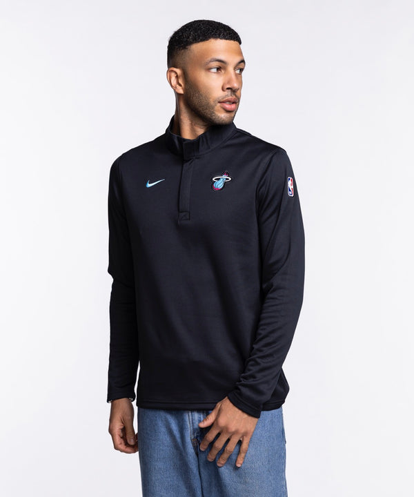 A man stands facing forward in a Nike Miami HEAT Original Vice Nights 1/4 Zip Jacket, featuring the team logo on the chest, paired with blue jeans. The black polyester pullover has a standard fit and is set against a plain white background.