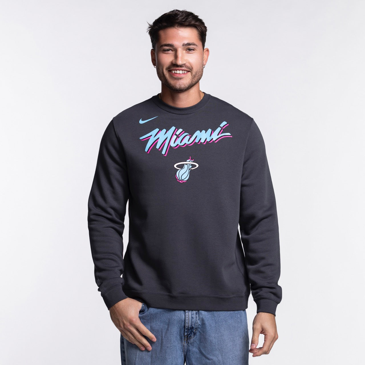 A man wearing a Nike Miami HEAT Original Vice Nights Crewneck sweatshirt with colorful Miami graphics stands smiling against a plain light background.