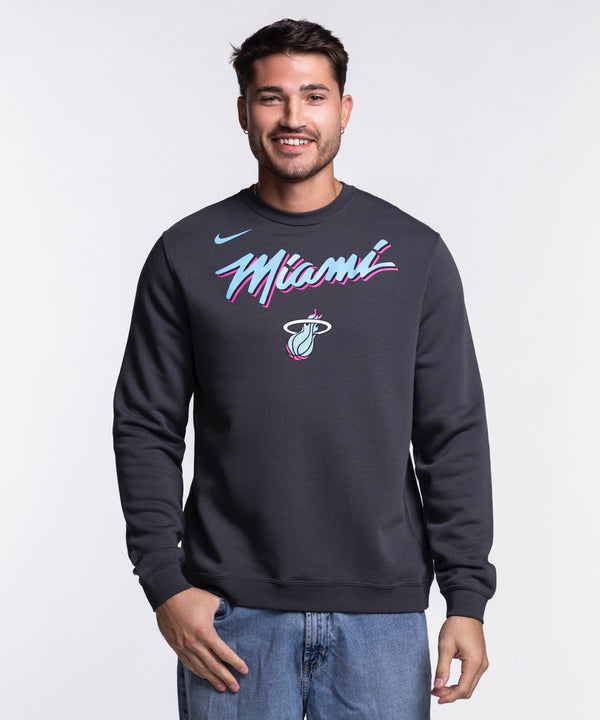 A man wearing a Nike Miami HEAT Original Vice Nights Crewneck sweatshirt with colorful Miami graphics stands smiling against a plain light background.