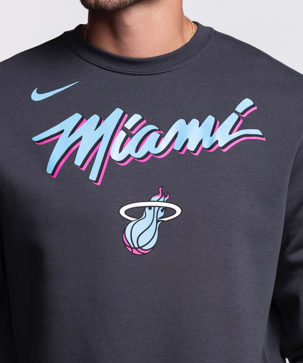 A man wearing a Nike Miami HEAT Original Vice Nights Crewneck sweatshirt with colorful Miami graphics stands smiling against a plain light background.