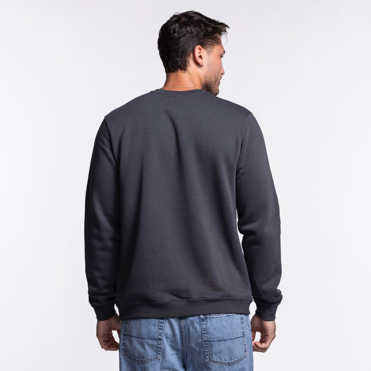 A man faces away from the camera, wearing a Nike Miami HEAT Original Vice Nights Crewneck sweatshirt in dark gray and light blue jeans, standing against a plain white background.
