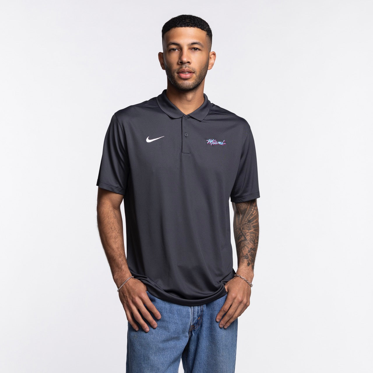 A man stands against a plain white background, wearing the Nike Miami HEAT Original Vice Nights Polo and blue jeans. The polyester shirt by Nike complements his relaxed stance, short hair, and forearm tattoos.