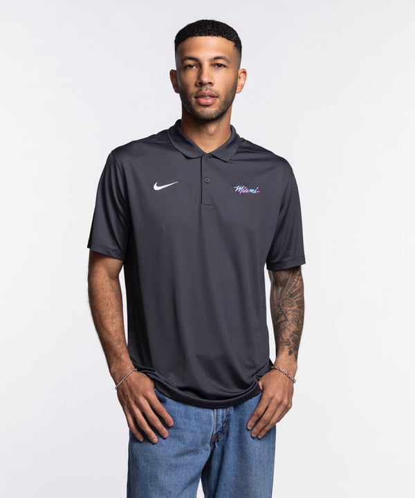 A man stands against a plain white background, wearing the Nike Miami HEAT Original Vice Nights Polo and blue jeans. The polyester shirt by Nike complements his relaxed stance, short hair, and forearm tattoos.