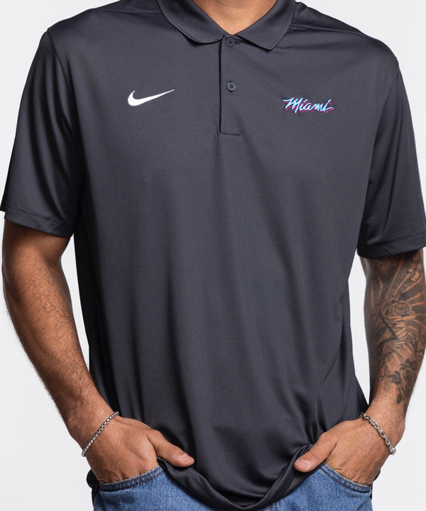 A man stands against a plain white background, wearing the Nike Miami HEAT Original Vice Nights Polo and blue jeans. The polyester shirt by Nike complements his relaxed stance, short hair, and forearm tattoos.