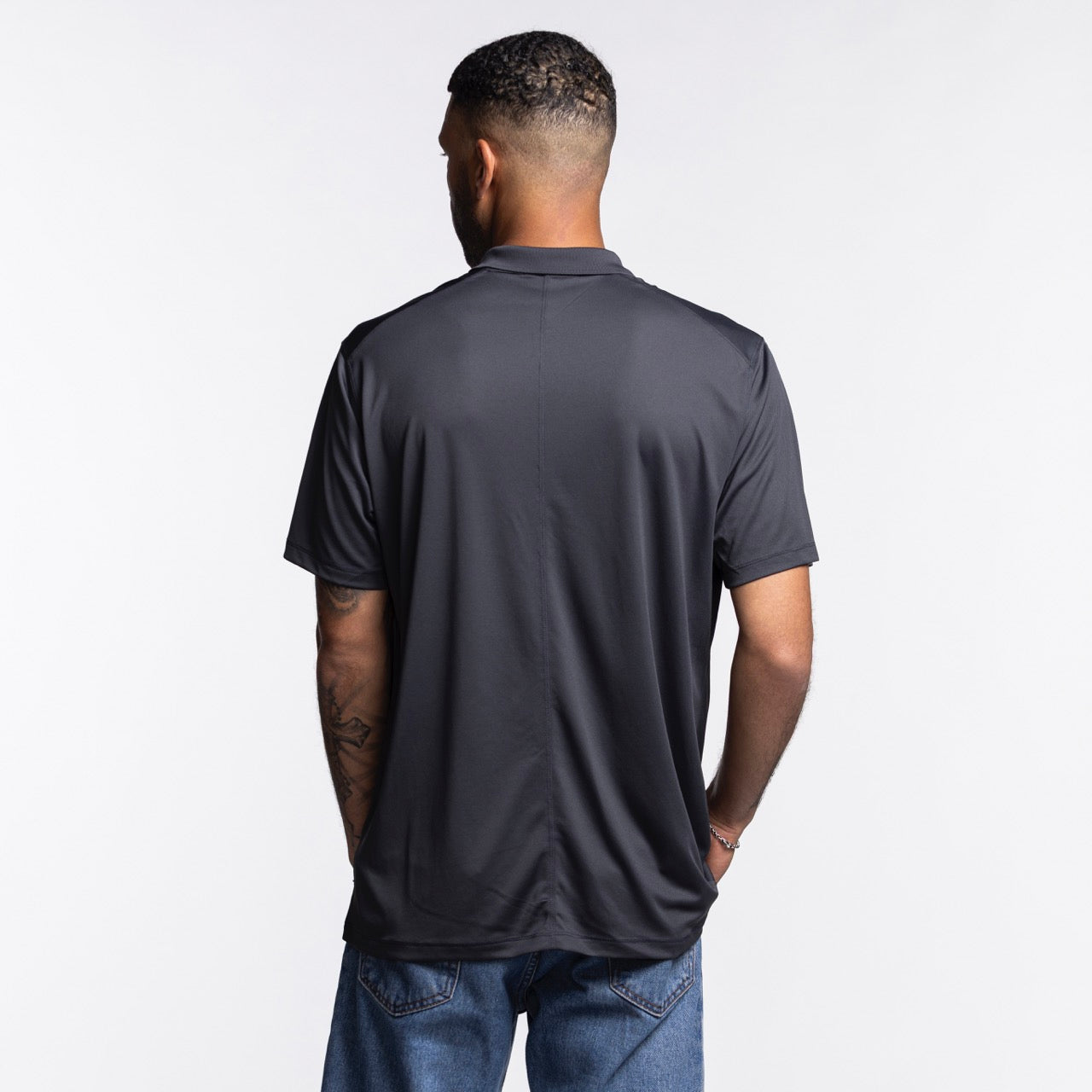 A man with short hair and visible arm tattoos stands with his back to the camera, wearing the Nike Miami HEAT Original Vice Nights Polo by Nike and blue jeans, set against a plain light background.