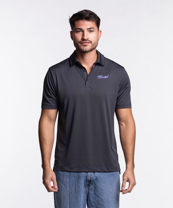 A man with short brown hair and a trimmed beard wears a Columbia Miami HEAT Original Vice Nights Drive Polo by Columbia Outdoor and blue jeans, standing against a plain white background.