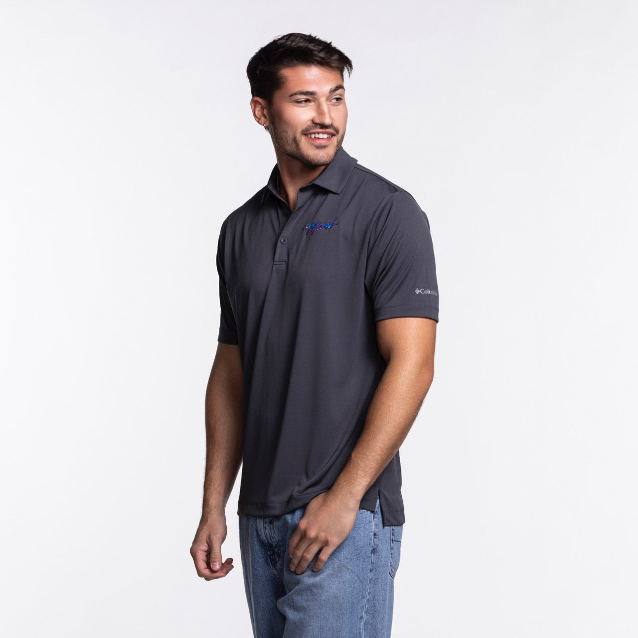 A man wearing a Columbia Outdoor Miami HEAT Original Vice Nights Drive Polo in dark gray and blue jeans stands looking left against a plain white background.