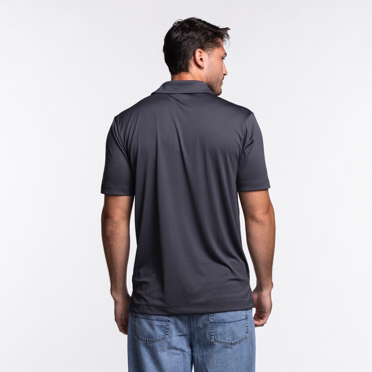 A man in a Columbia Outdoor Miami HEAT Original Vice Nights Drive Polo, standard fit with short sleeves in dark gray, and blue jeans stands facing away from the camera against a plain white background.
