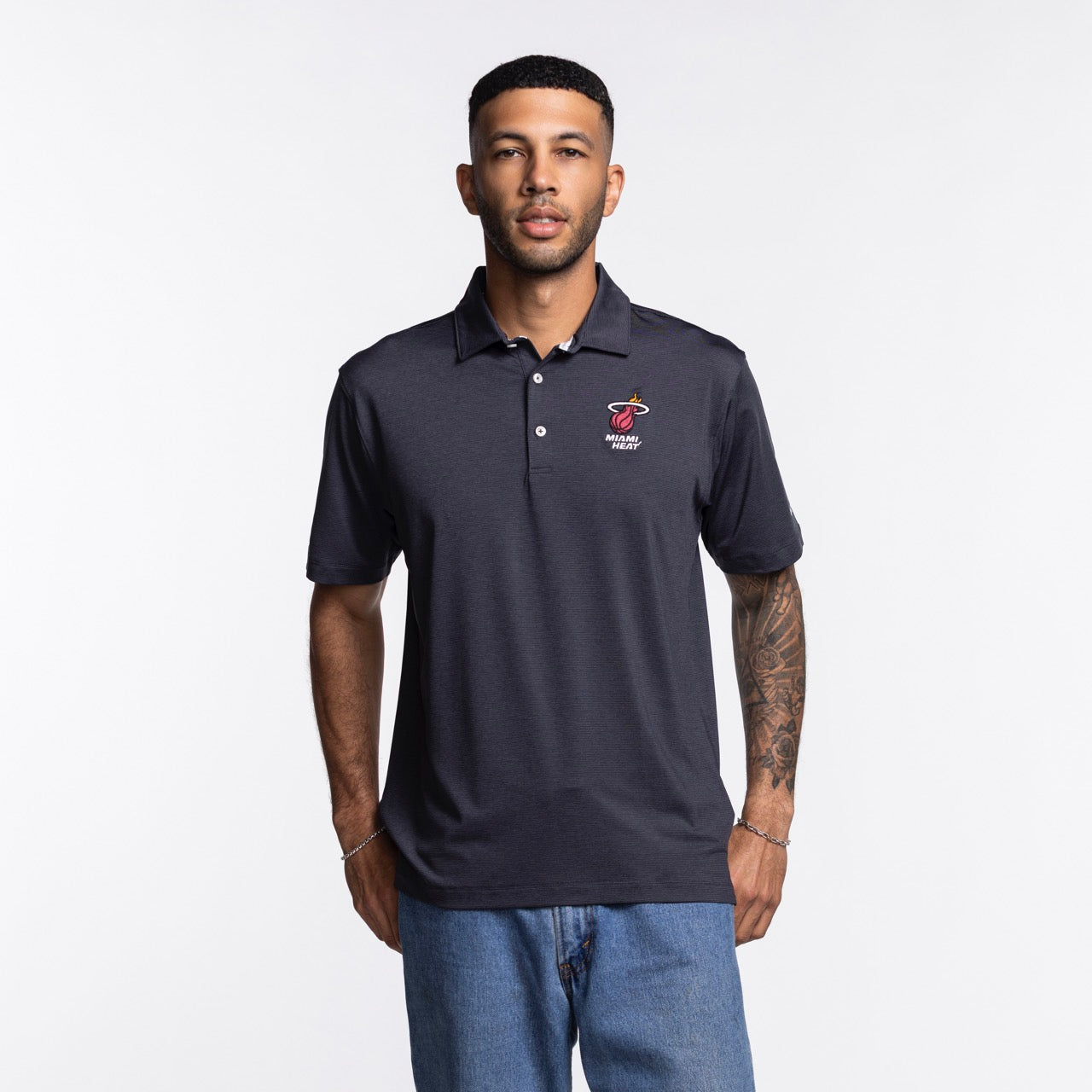 A man wearing a Columbia Miami HEAT Logo Grey Polo by Columbia Outdoor and light blue jeans stands against a plain white background, facing the camera with a neutral expression. The polo is polyester-spandex and machine washable.