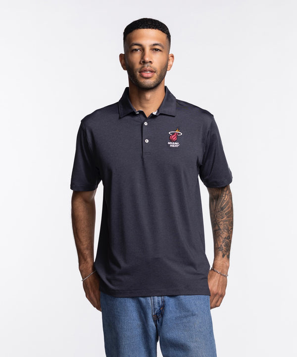A man wearing a Columbia Miami HEAT Logo Grey Polo by Columbia Outdoor and light blue jeans stands against a plain white background, facing the camera with a neutral expression. The polo is polyester-spandex and machine washable.
