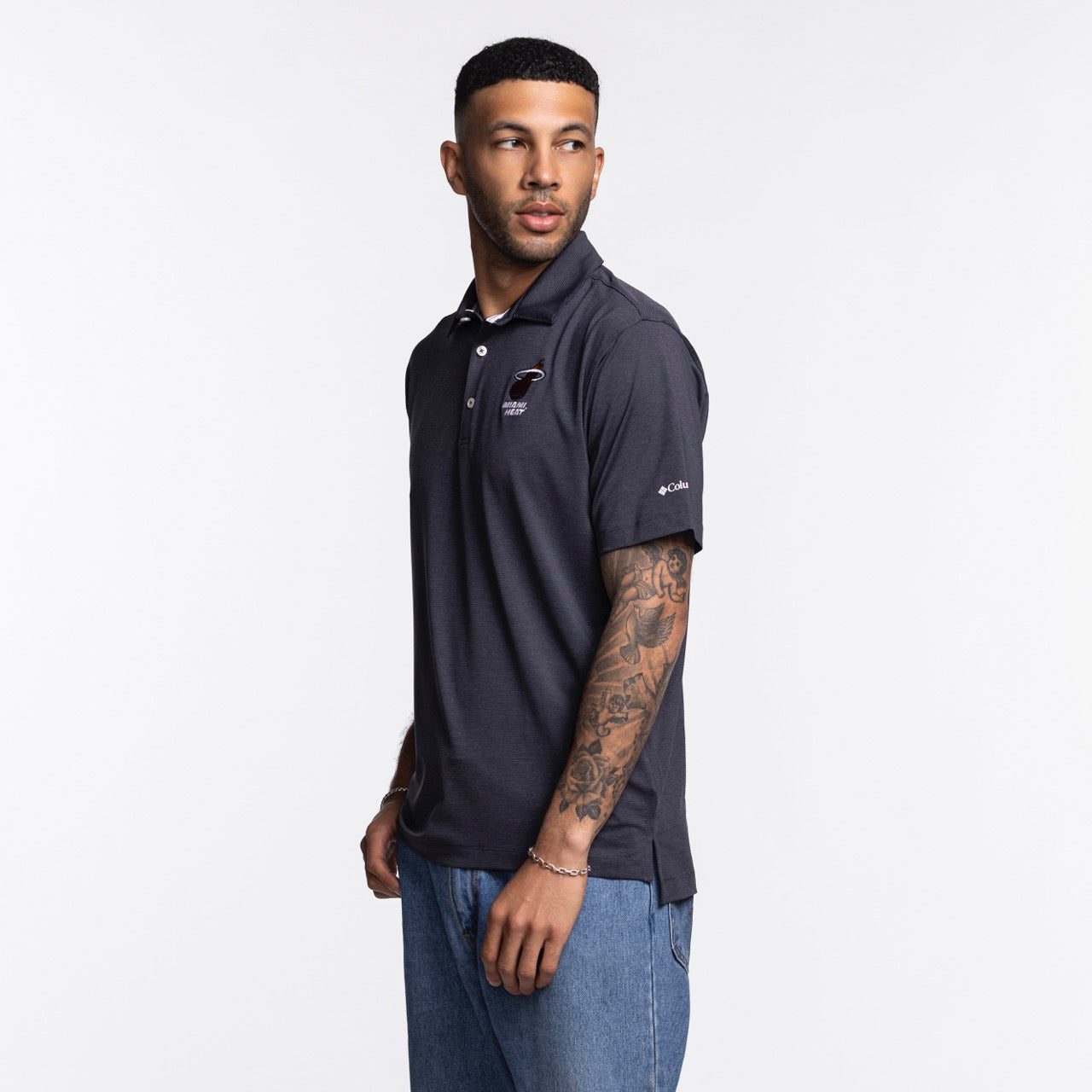 A man with short dark hair and tattoos on his left arm models the Columbia Miami HEAT Logo Grey Polo by Columbia Outdoor with blue jeans, standing against a plain white background and looking slightly to his left.