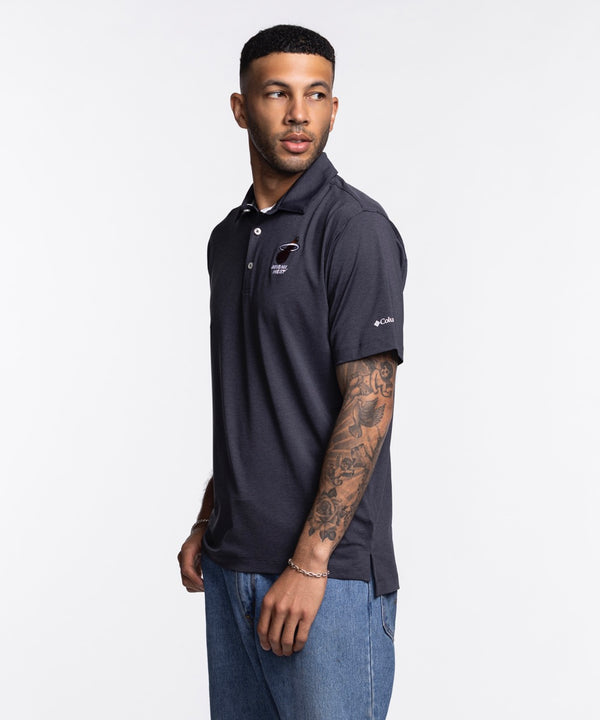A man wearing a Columbia Miami HEAT Logo Grey Polo by Columbia Outdoor and light blue jeans stands against a plain white background, facing the camera with a neutral expression. The polo is polyester-spandex and machine washable.
