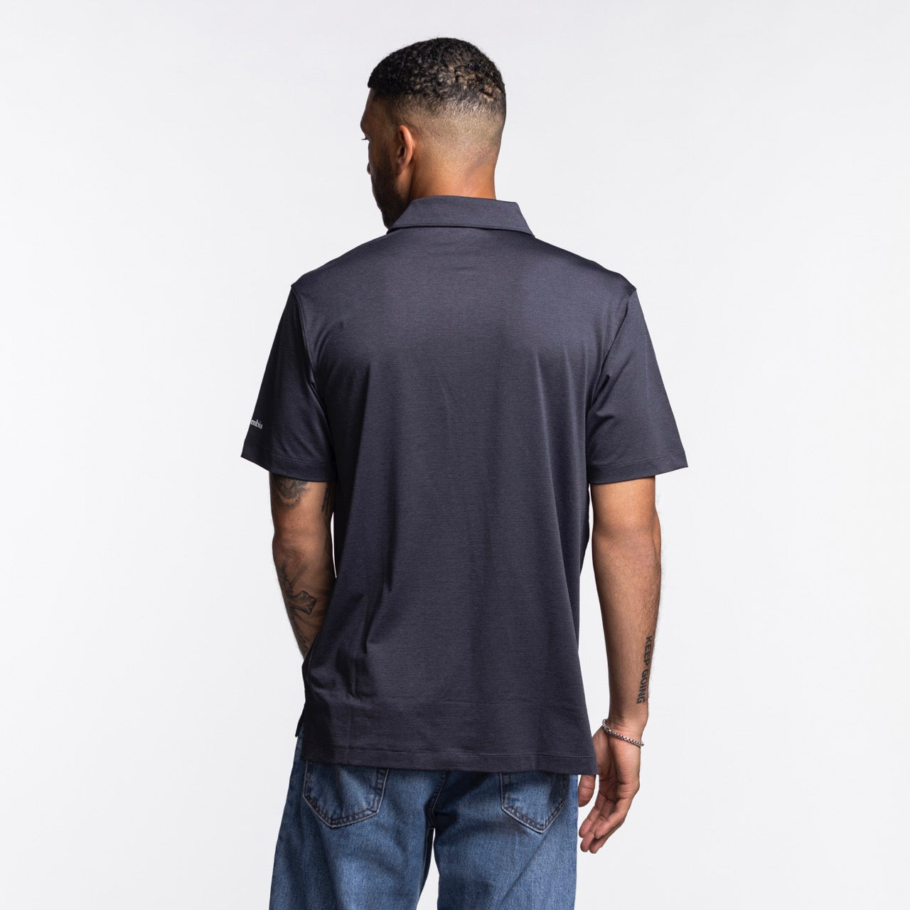 A man with short hair and a trimmed beard stands with his back to the camera, wearing the Columbia Miami HEAT Logo Grey Polo by Columbia Outdoor and blue jeans against a plain white background.
