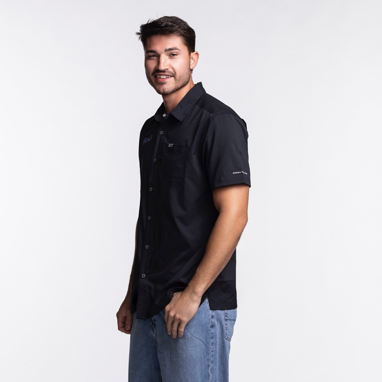 A young man with short dark hair, slight beard, and mustache smiles while wearing the Columbia Miami HEAT Original Vice Nights Slack Tide Shirt by Columbia Outdoor and light blue jeans, standing against a plain white background.