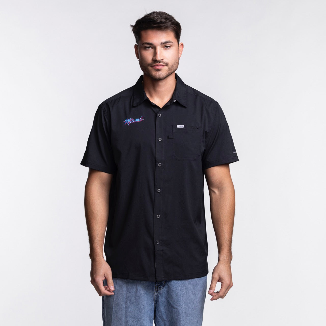 A man with short dark hair and a beard wears the Columbia Miami HEAT Original Vice Nights Slack Tide Shirt by Columbia Outdoor and light blue jeans, standing against a plain white background.