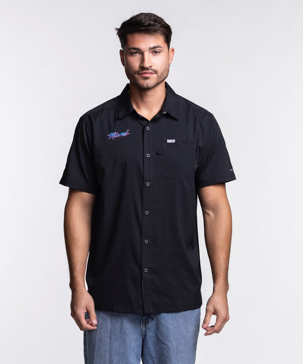 A man with short dark hair and a beard wears the Columbia Miami HEAT Original Vice Nights Slack Tide Shirt by Columbia Outdoor and light blue jeans, standing against a plain white background.