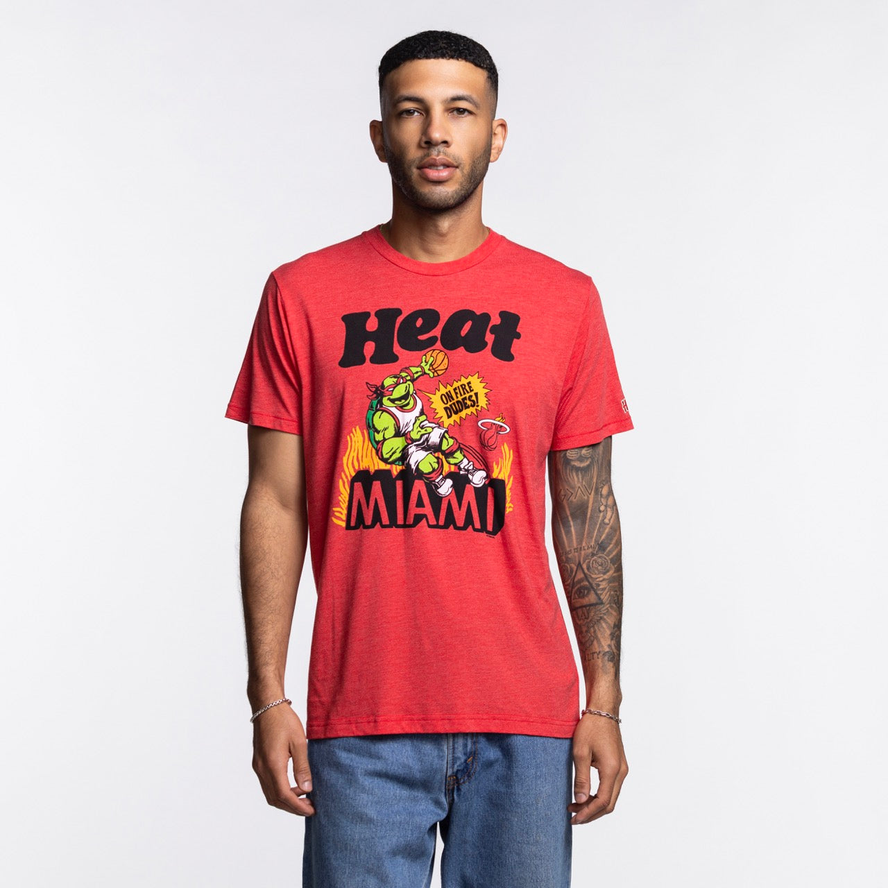 A man with short hair, a trimmed beard, and a left arm tattoo wears the Homage Miami HEAT Red TMNT Tee by Homage—featuring a colorful graphic and Heat Miami text. The standard-fit tee pairs well with his blue jeans. Made from polyester cotton rayon.