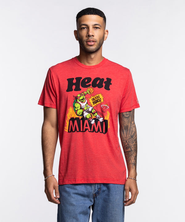 A man with short hair, a trimmed beard, and a left arm tattoo wears the Homage Miami HEAT Red TMNT Tee by Homage—featuring a colorful graphic and Heat Miami text. The standard-fit tee pairs well with his blue jeans. Made from polyester cotton rayon.