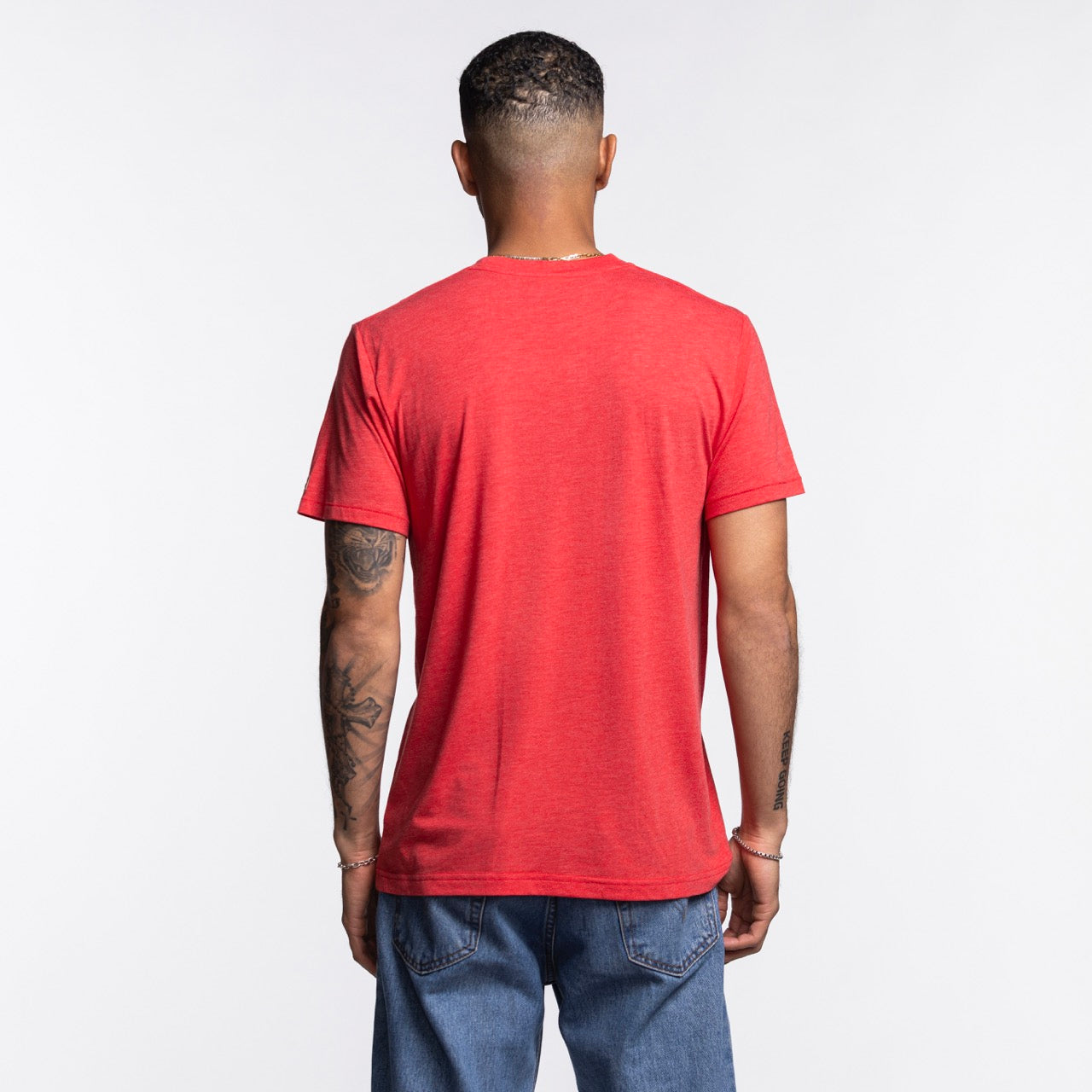 A man stands with his back to the camera, wearing the Homage Miami HEAT Red TMNT Tee by Homage and blue jeans. His tattooed arms rest at his sides. The soft tee is made from a polyester, cotton, and rayon blend against a plain light background.