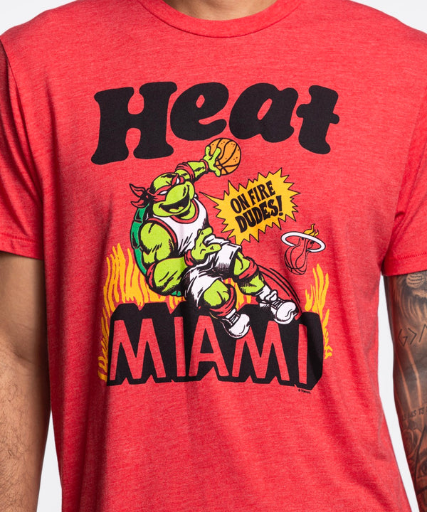 A man with short hair, a trimmed beard, and a left arm tattoo wears the Homage Miami HEAT Red TMNT Tee by Homage—featuring a colorful graphic and Heat Miami text. The standard-fit tee pairs well with his blue jeans. Made from polyester cotton rayon.