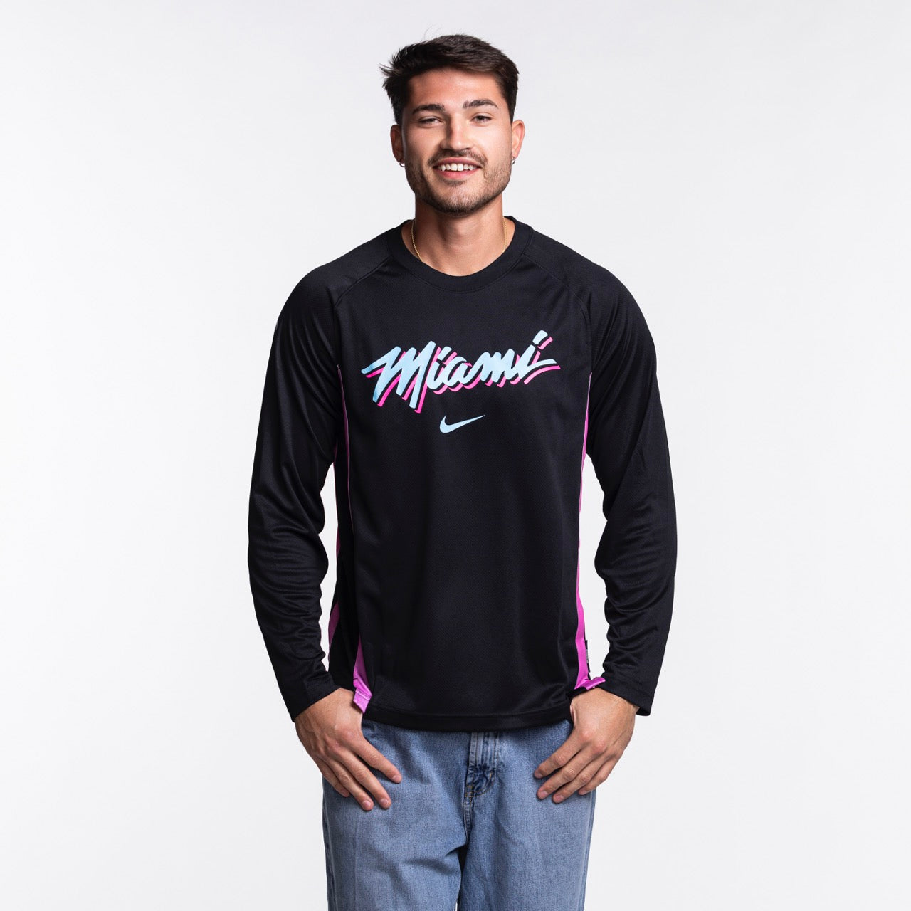 A man with short dark hair and light facial hair smiles against a plain light background, wearing a Nike Miami HEAT Original Vice Nights Shooting Shirt in black and blue jeans; wash with like colors recommended.