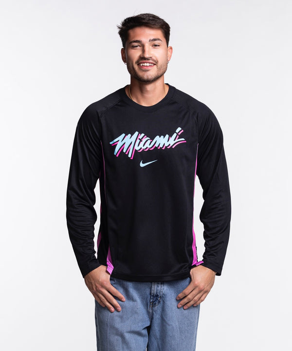 A man with short dark hair and light facial hair smiles against a plain light background, wearing a Nike Miami HEAT Original Vice Nights Shooting Shirt in black and blue jeans; wash with like colors recommended.