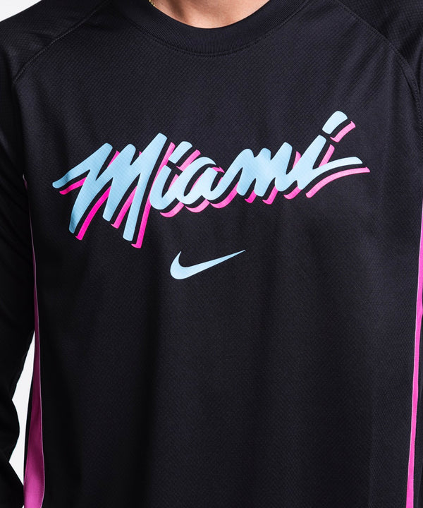 A man with short dark hair and light facial hair smiles against a plain light background, wearing a Nike Miami HEAT Original Vice Nights Shooting Shirt in black and blue jeans; wash with like colors recommended.