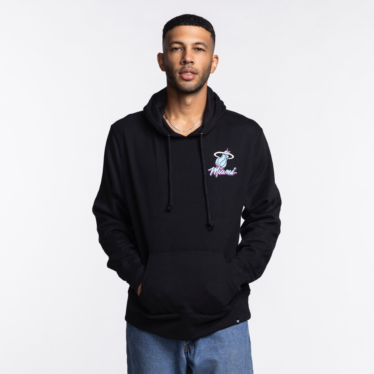 A man faces forward in a '47 Brand Miami HEAT Original Vice Nights Hoodie, featuring the basketball logo. The black cotton-polyester hoodie is machine washable and paired with light blue jeans against a plain white background.