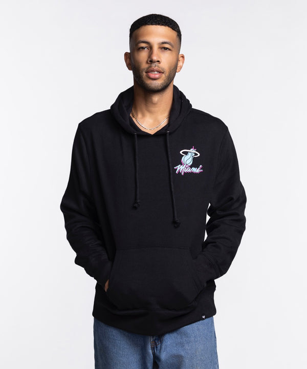 A man faces forward in a '47 Brand Miami HEAT Original Vice Nights Hoodie, featuring the basketball logo. The black cotton-polyester hoodie is machine washable and paired with light blue jeans against a plain white background.