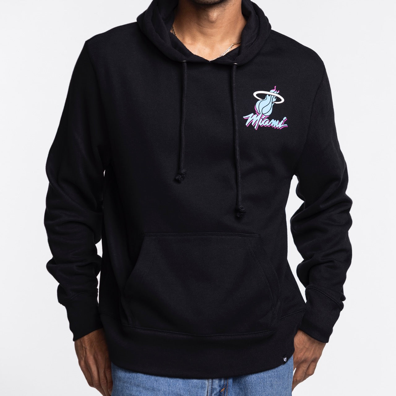 A person wears the '47 Brand Miami HEAT Original Vice Nights Hoodie in black (cotton-poly blend) with a pink and blue Miami Heat logo on the chest, paired with blue jeans against a white background; machine washable.