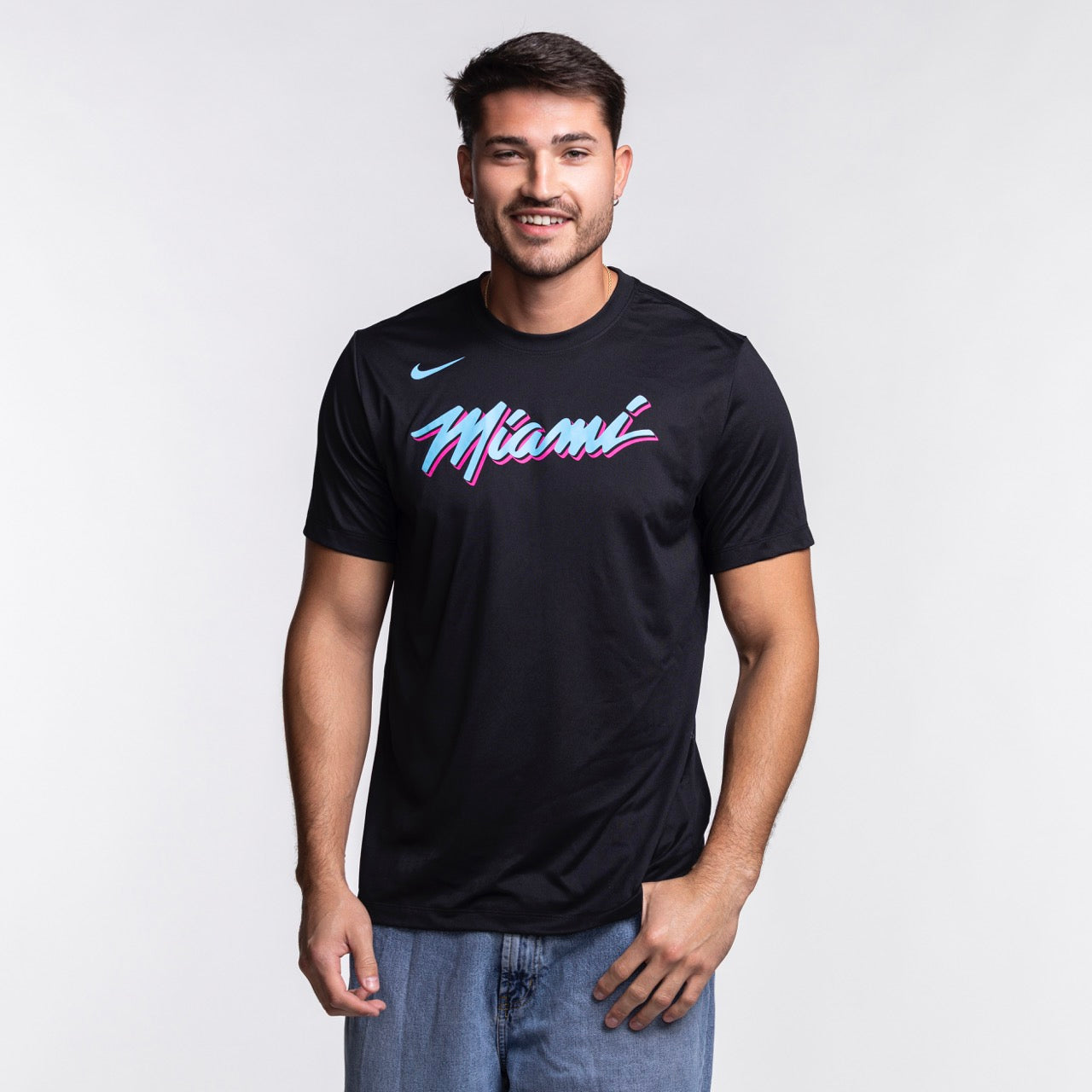 A man with short dark hair and a beard smiles in a Nike Miami HEAT Original Vice Nights Legend Tee—black with colorful “Miami” text—by Nike, paired with blue jeans, standing against a plain light background.