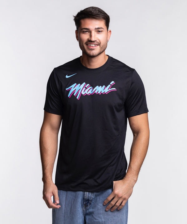 A man with short dark hair and a beard smiles in a Nike Miami HEAT Original Vice Nights Legend Tee—black with colorful “Miami” text—by Nike, paired with blue jeans, standing against a plain light background.