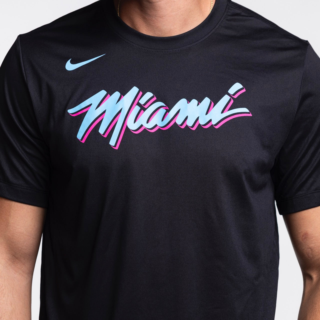 A person wears the Nike Miami HEAT Original Vice Nights Legend Tee—black polyester, standard fit, featuring “Miami” in blue, pink, and white script across the chest. Only the torso is visible; wash with like colors.