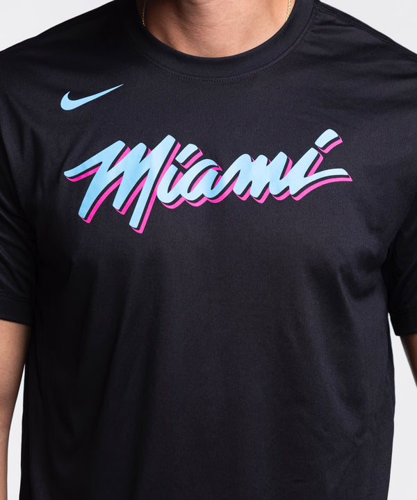 A man with short dark hair and a beard smiles in a Nike Miami HEAT Original Vice Nights Legend Tee—black with colorful “Miami” text—by Nike, paired with blue jeans, standing against a plain light background.