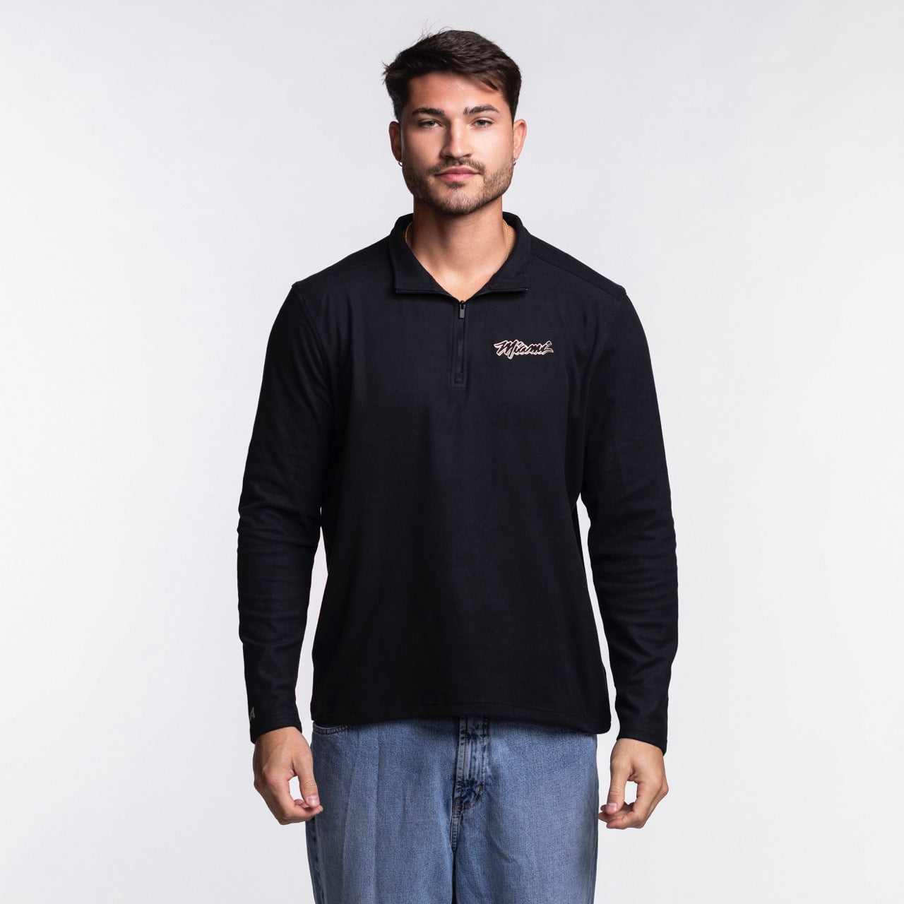 A man with short dark hair and light facial hair wears the Antigua Miami HEAT Original Vice Nights 1/4 Zip Jacket by Antigua, featuring a black polyester-spandex blend for comfort and standard fit, paired with light blue jeans. Machine wash cold.