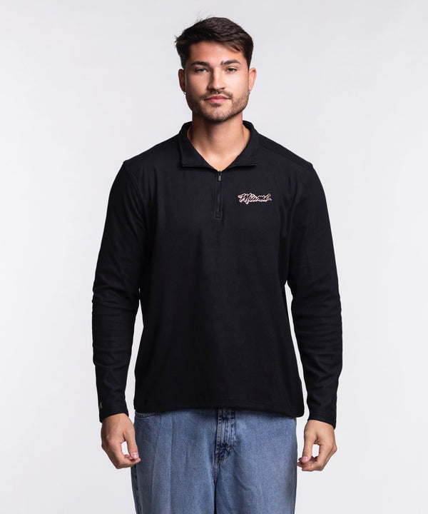 A man with short dark hair and light facial hair wears the Antigua Miami HEAT Original Vice Nights 1/4 Zip Jacket by Antigua, featuring a black polyester-spandex blend for comfort and standard fit, paired with light blue jeans. Machine wash cold.