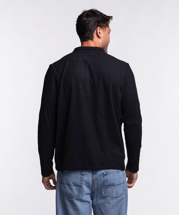 A man with short dark hair and light facial hair wears the Antigua Miami HEAT Original Vice Nights 1/4 Zip Jacket by Antigua, featuring a black polyester-spandex blend for comfort and standard fit, paired with light blue jeans. Machine wash cold.