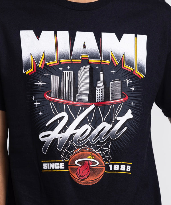 A man stands against a white background wearing the Miami HEAT Retro Net Tee by Item Of The Game, featuring a city skyline graphic. He faces forward with his hands in his pockets. This standard-fit cotton shirt is machine washable.