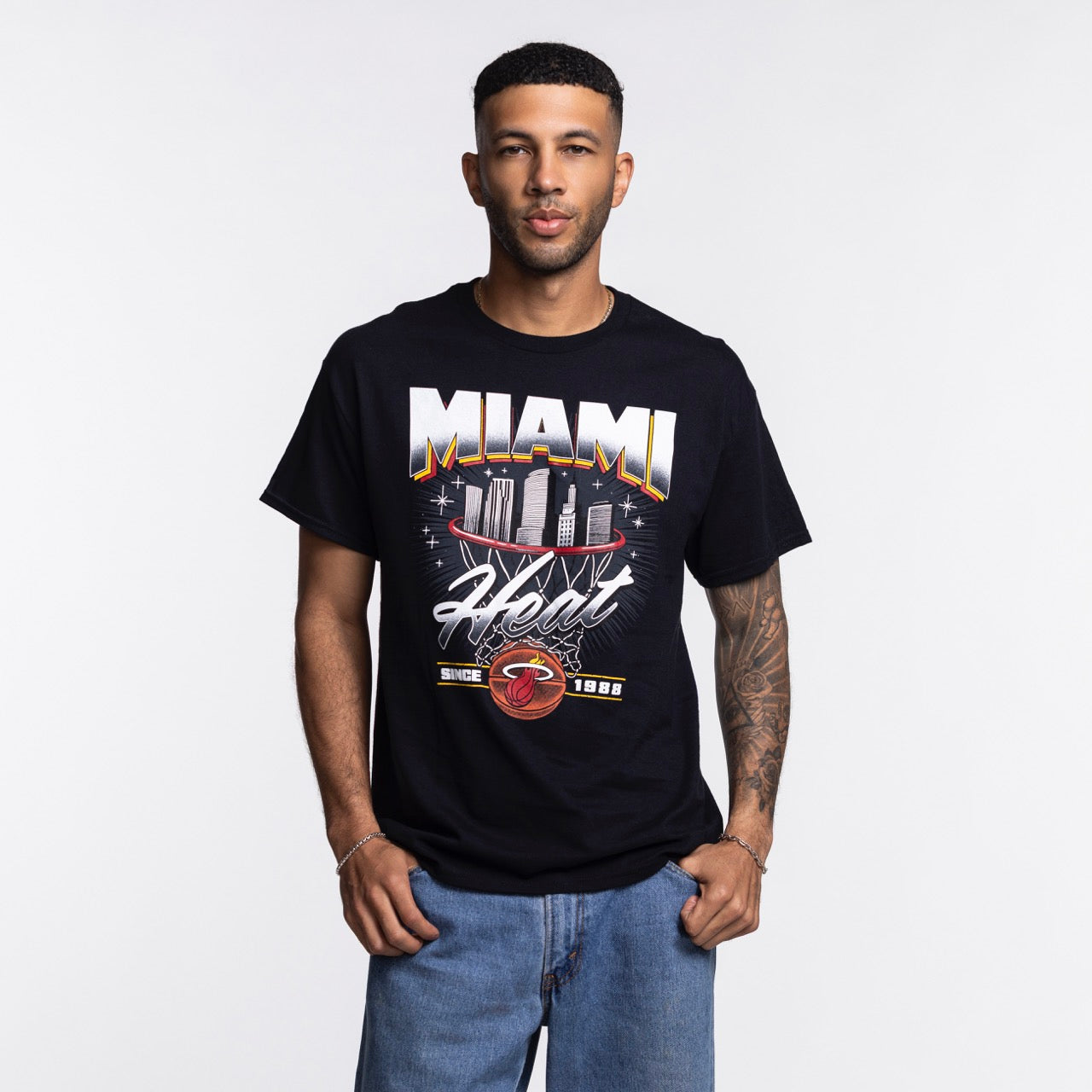A man stands against a white background wearing the Miami HEAT Retro Net Tee by Item Of The Game, featuring a city skyline graphic. He faces forward with his hands in his pockets. This standard-fit cotton shirt is machine washable.