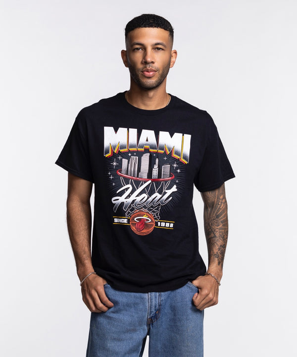 A man stands against a white background wearing the Miami HEAT Retro Net Tee by Item Of The Game, featuring a city skyline graphic. He faces forward with his hands in his pockets. This standard-fit cotton shirt is machine washable.