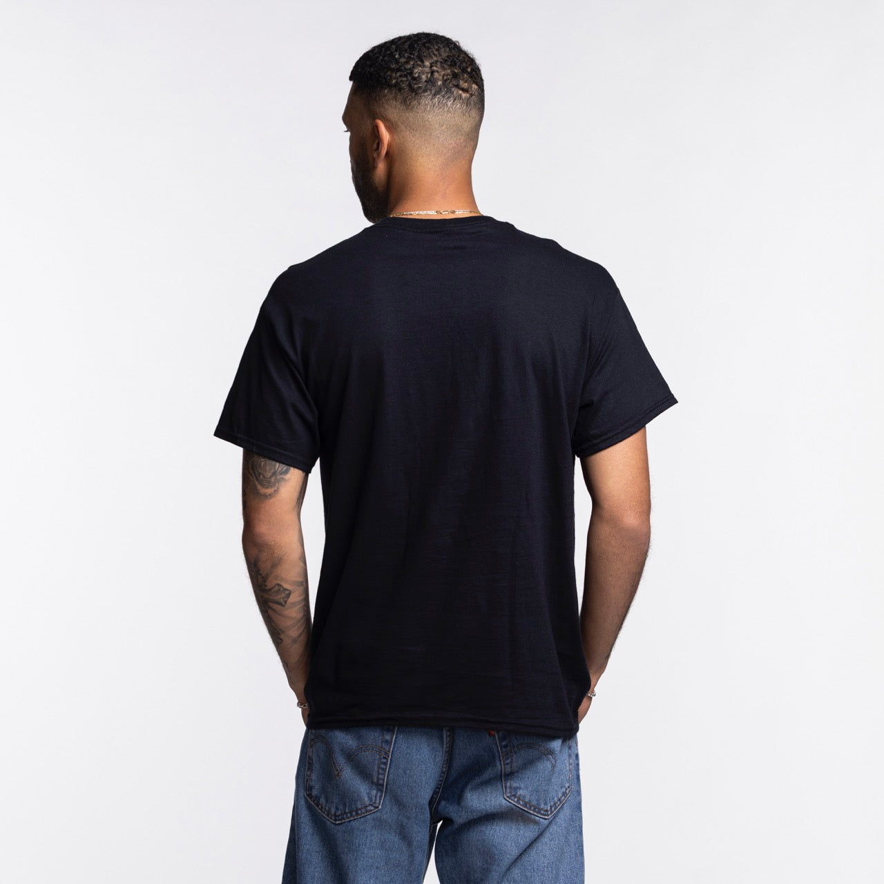 A man with short hair and tattoos on his left arm faces away from the camera, wearing the Item Of The Game Miami HEAT Retro Net Tee and blue jeans against a plain white background.