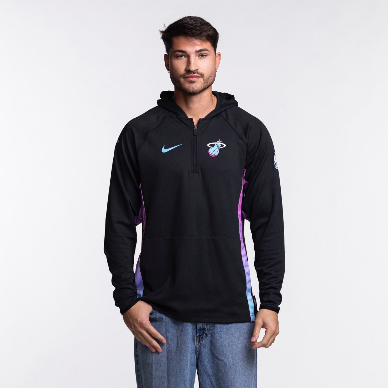 A man faces forward in a Nike Miami HEAT Original Vice Nights 1/2 Zip Hoodie, featuring blue and pink accents, paired with blue jeans. He has a neutral expression and stands against a plain white background.